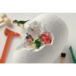 A white dinosaur hatching egg toy with a cracked shell, shown open with small dinosaur figurines emerging inside. Surrounding the egg are toy tools including a hammer, chisel, brush, and small dinosaur figures on a white background.