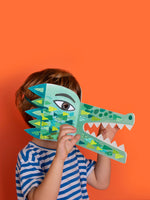 Child holding a green paper crocodile mask against an orange background