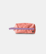 A peach-pink nylon pouch with a lavender purple horizontal strap across the front and a small black circular logo patch on the lower right, shown on a light gray background.