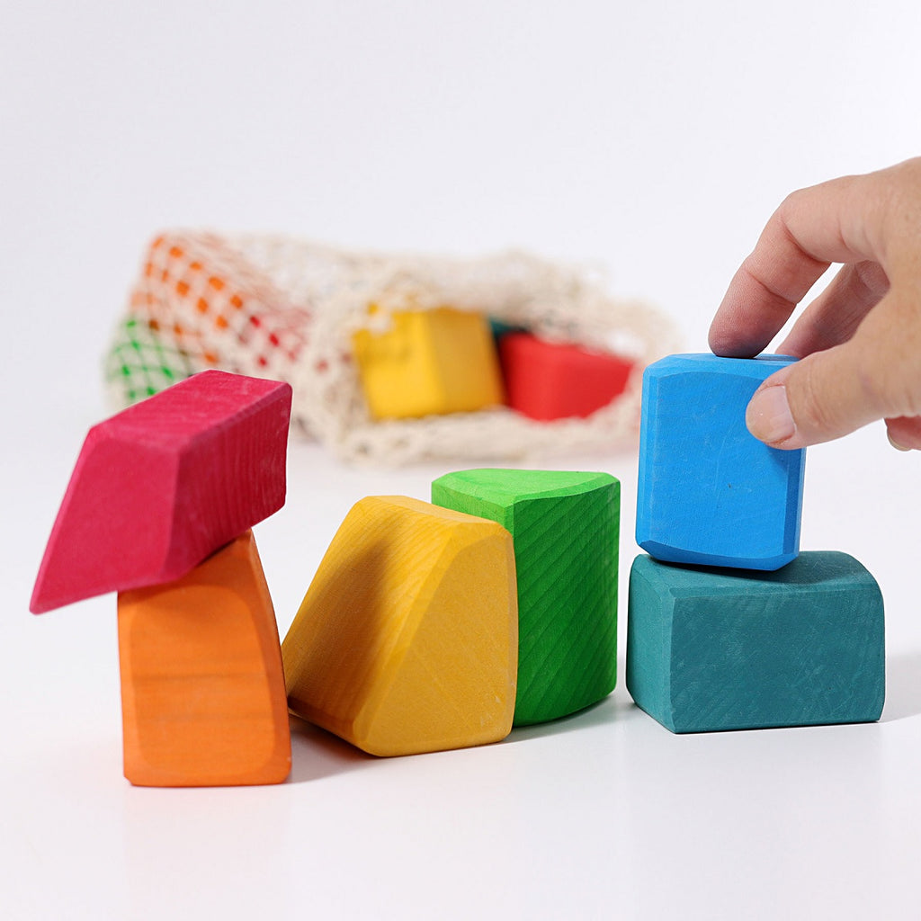 a hand stacking a blue wooden block onto other colourful blocks in the foreground. The rest of the blocks are in a net in the background. In a white setting.