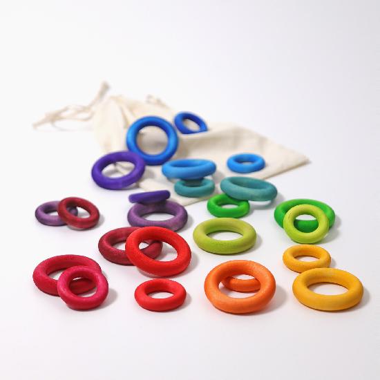 On a white background, two sizes of wooden rings spread out in rainbow colours. A cotton bag is underneath them.