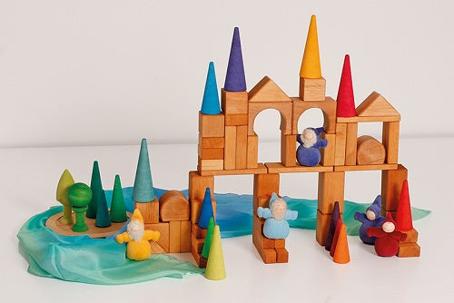 in a white room, wooden building blocks coupled with rainbow coloured cones arranged in a structure with a blue piece of silk arranged at the bottom. Plush gnomes are included in the structure.