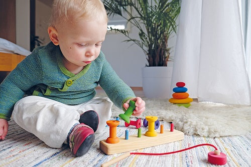 an interior scene, a toddler sitting on the floor placing a green bobbin on a peg attached to a wooden board. 