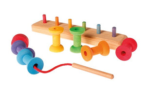 on a white background, a natural wooden board with 8 rainbow coloured pegs. Matching coloured spools are in front of the board. A wooden needle attached to a red string is in the foreground. Two bobbins are threaded onto it.