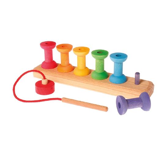 On a white background, 8 rainbow coloured bobbins on a board with a wooden needle attached to a red string ended with a red cap in the foreground. One purple bobbin is in front of the board revealing a purple peg that the bobbin can be placed on.