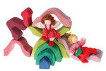 Pieces of a wooden Lotus flower stacker in red, pink, purples, greens and blue with dolls using pieces as beds and slides on a white background
