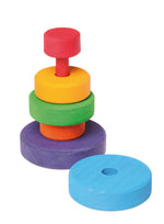 On a white background, a small wooden stacking tower with rings in rainbow colours. The rings are stacked outside of size and colour order in a creative fashion. One blue ring rests next to the tower