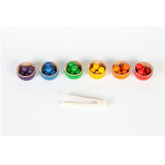 A series of 6 bowls in rainbow order filled with coordinating coloured acorns on a white background. A set of tongs are in the foreground