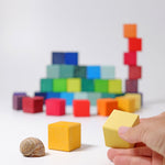 in a white space, a hand holding a yellow cube in the foreground. Also in the foreground, another yellow cube and a snail shell. In the background, a pile of wooden rainbow coloured cubes