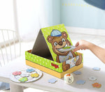 interior shot, a child's hand using a flat spoon to put food into an illustrated bears mouth on the box that is on the table.