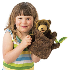 A little girl with brown hair and bangs in a striped tank top with her hand in the bottom of a brown, plush bear cub in a soft tree stump with two green leaves on a white background.