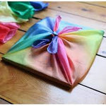 on a wooden floor, a flat square package wrapped in a gradient rainbow silk tied on the top.