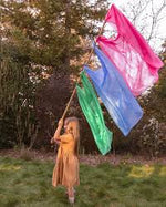 a girl in a forest holding a long stick with playsilks tied to it like a flag
